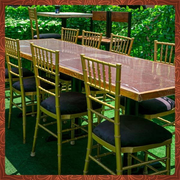 Long dining table with gold bamboo-style chairs and black cushions on a green turf patio.