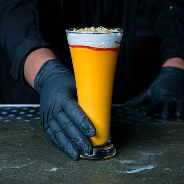 A gloved hand holds a tall glass of orange mango lassi, topped with cream and nuts.