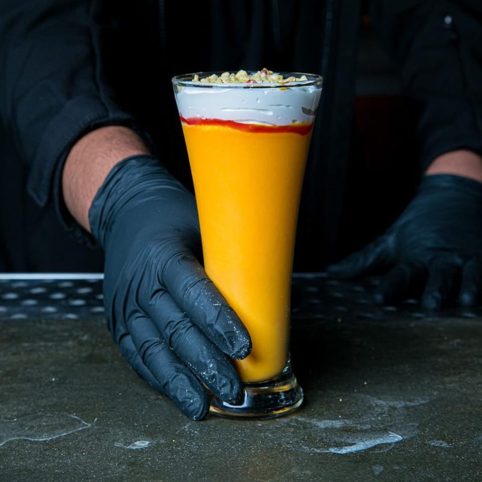 A gloved hand holds a tall glass of orange mango lassi, topped with cream and nuts.