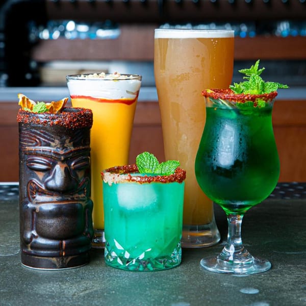Four vibrant drinks, including a tiki cocktail, a mango lassi, a beer, and two green cocktails.