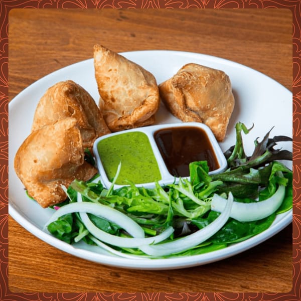 Four golden-brown samosas with green and brown chutneys, and a fresh salad on a white plate.