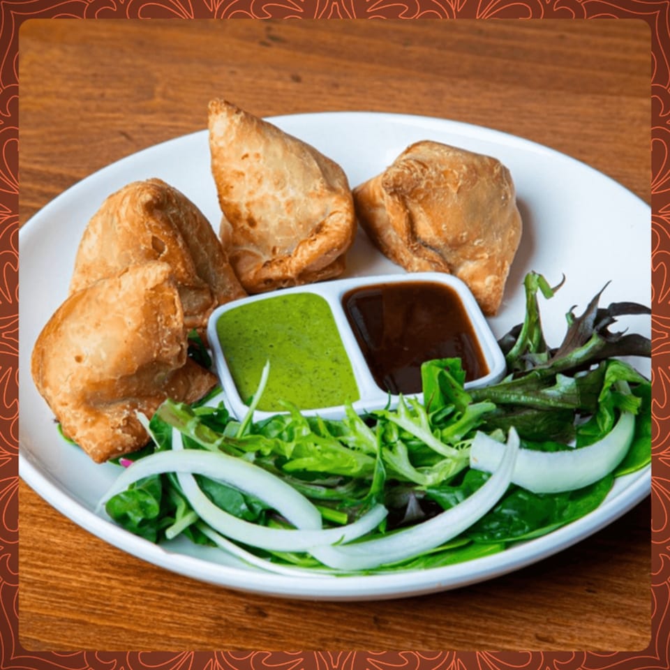 Four golden-brown samosas with green and brown chutneys, and a fresh salad on a white plate.
